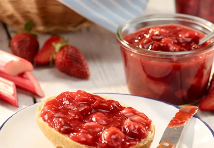 Ein Glas dunkle Rhabarber‑Erdbeer‑Konfitüre steht auf einem Brett, umgeben von Erdbeeren, Rhabarber und einer Zitronenspalte.