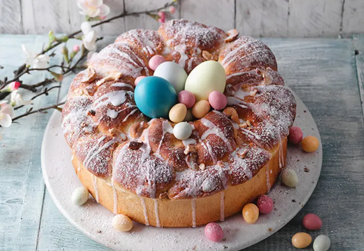 Ein ringförmiger Hefekranz aus Dinkelteig mit Cashew-Frischkäse-Füllung, orangefarbener Glasur und bunten Zucker-Ostereiern