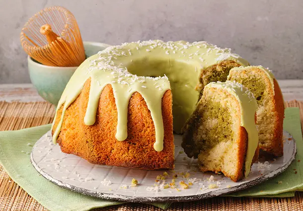 Ein Gugelhupf mit hellem Kokos- und grünem Matchateig, marmoriert, mit hellgrünem Matcha-Zuckerguss überzogen und mit Kokosraspeln bestreut