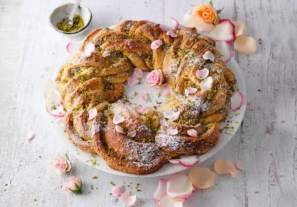 Ein geflochtener Hefekranz mit Pistazienfüllung und kandierten Rosenblüten ist leicht mit Puderzucker bestäubt