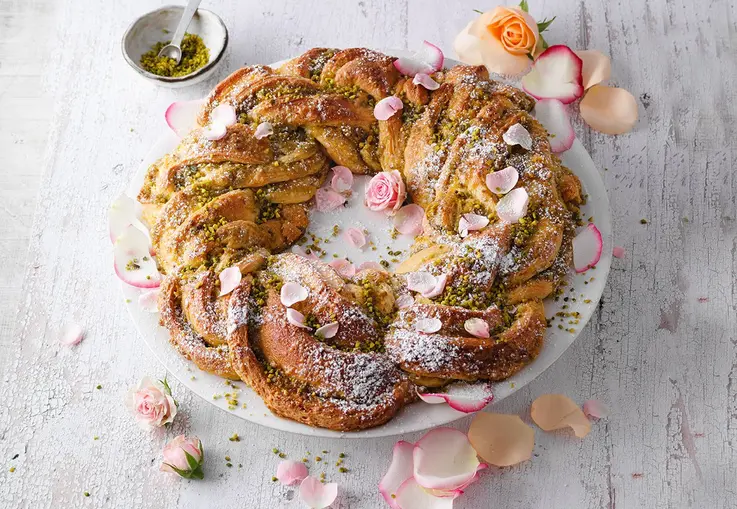 Ein geflochtener Hefekranz mit Pistazienfüllung und kandierten Rosenblüten ist leicht mit Puderzucker bestäubt