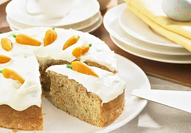 Ein runder Karottenkuchen mit Haselnüssen, bedeckt mit cremigem Frischkäse‑Frosting und kleinen Marzipanmöhren; ein Stück ist herausgeschnitten