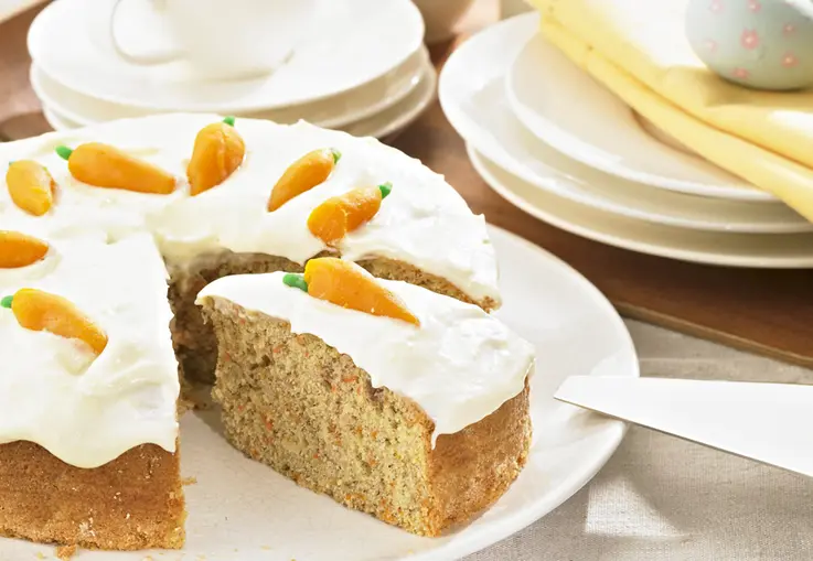 Ein runder Karottenkuchen mit Haselnüssen, bedeckt mit cremigem Frischkäse‑Frosting und kleinen Marzipanmöhren; ein Stück ist herausgeschnitten