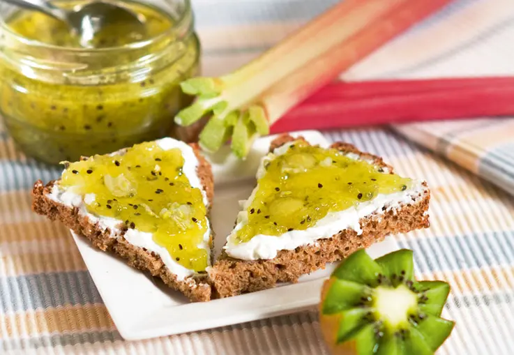 Toastscheiben mit grüner Rhabarber‑Stachelbeer‑Kiwi‑Konfitüre liegen neben einem Glas des exotischen Aufstrichs und frischem Obst.