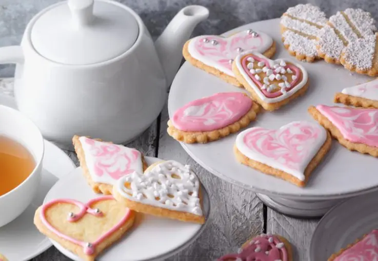 Verschieden geformte Butterplätzchen aus Mürbeteig, mit rosafarbenem und weißem Zuckerguss und Zuckerperlen dekoriert, auf einer Platte angerichtet