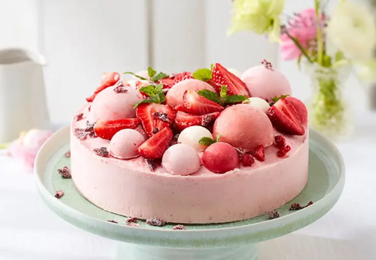 Ein gefrorener Erdbeer‑Rhabarber‑Cheesecake auf einem Teller, mit roten Fruchtschichten und Zitronenmelisse garniert.