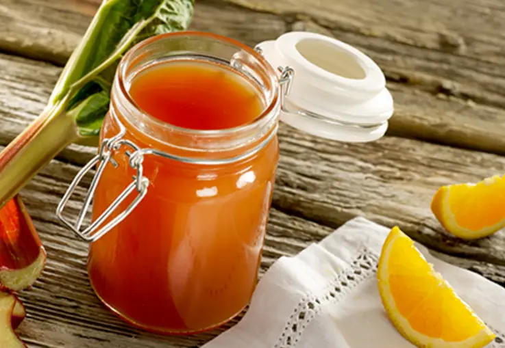 Ein Glas rosafarbenes Rhabarber‑Orangen‑Gelee steht neben Orangenscheiben und frischen Rhabarberstücken.
