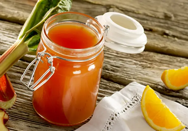 Ein Glas rosafarbenes Rhabarber‑Orangen‑Gelee steht neben Orangenscheiben und frischen Rhabarberstücken.