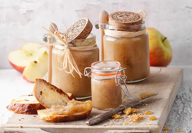 Zwei kleine Gläser Apfel-Sesam-Aufstrich stehen auf einem Brett; daneben Brotscheiben und rote Äpfel