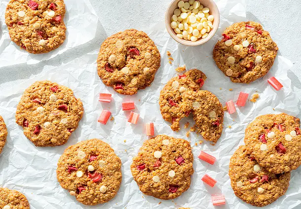Mehrere runde Cookies mit Rhabarberstücken und weißen Schokostückchen liegen auf Backpapier