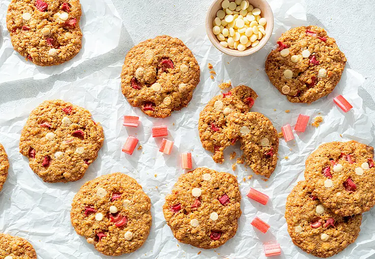 Mehrere runde Cookies mit Rhabarberstücken und weißen Schokostückchen liegen auf Backpapier