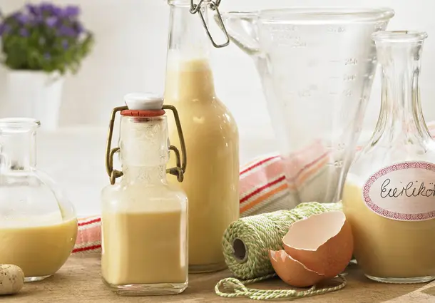 Mehrere Flaschen und Gläser mit cremigem Eierlikör aus Eigelb, Zucker, Kondensmilch und Rum; daneben ein zerbrochenes Ei und Küchenschnur