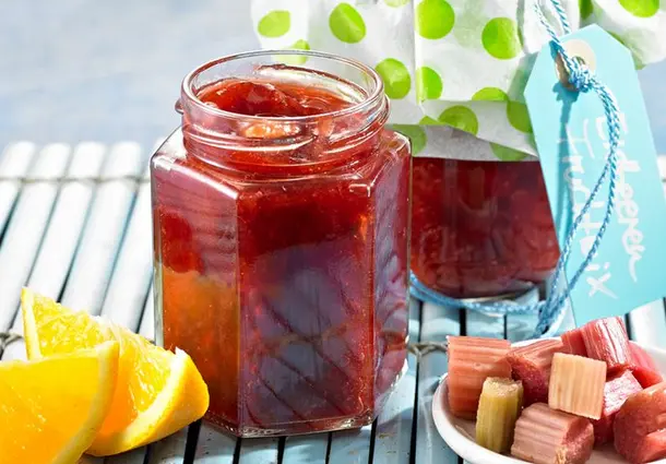 Gläser mit rotem Erdbeer‑Rhabarber‑Orangen‑Mix stehen auf einem Tisch, daneben liegen Orangenhälften und Rhabarberstücke.