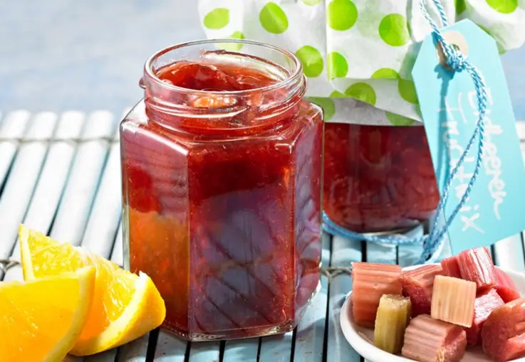 Gläser mit rotem Erdbeer‑Rhabarber‑Orangen‑Mix stehen auf einem Tisch, daneben liegen Orangenhälften und Rhabarberstücke.