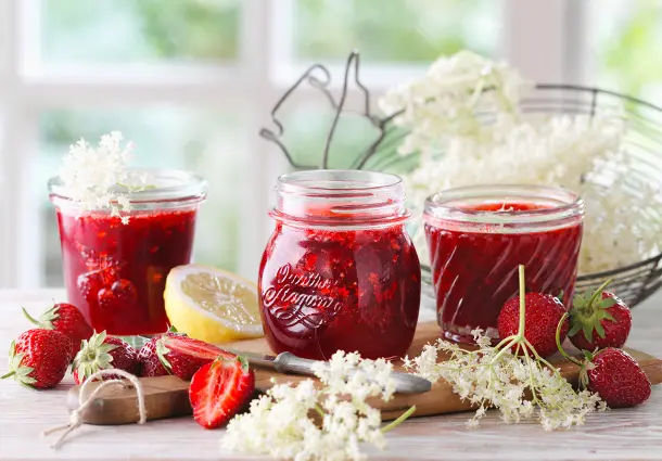 Gläser mit leuchtend roter Erdbeerkonfitüre, verfeinert mit kleinen weißen Holunderblüten.