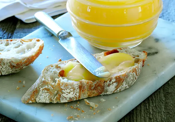 Ein Glas und eine Schüssel mit cremigem Lemon Curd aus Zitronensaft, Butter, Zucker und Eiern, daneben eine bestrichene Brotscheibe