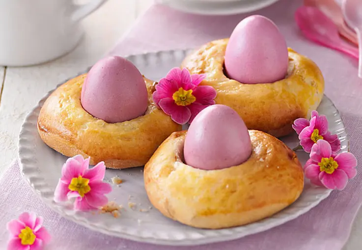 Kleine runde Brioche-Nester mit grobem Zucker und gehackten Pinienkernen, in der Mitte jeweils ein bunt gefärbtes Osterei, dekoriert mit Blüten