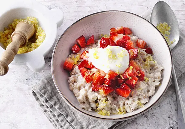Cremiges Mehrkorn‑Porridge in einer Schale, getoppt mit Erdbeerwürfeln, Joghurtcreme und Orangenzucker, daneben ein Mörser