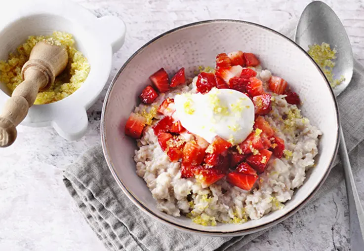 Cremiges Mehrkorn‑Porridge in einer Schale, getoppt mit Erdbeerwürfeln, Joghurtcreme und Orangenzucker, daneben ein Mörser