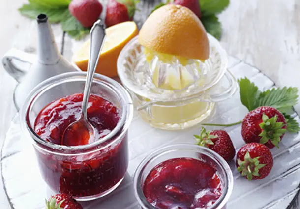 Glas mit dunkelroter Erdbeer‑Orangen‑Konfitüre, daneben frisch gepresste Orange und Erdbeeren