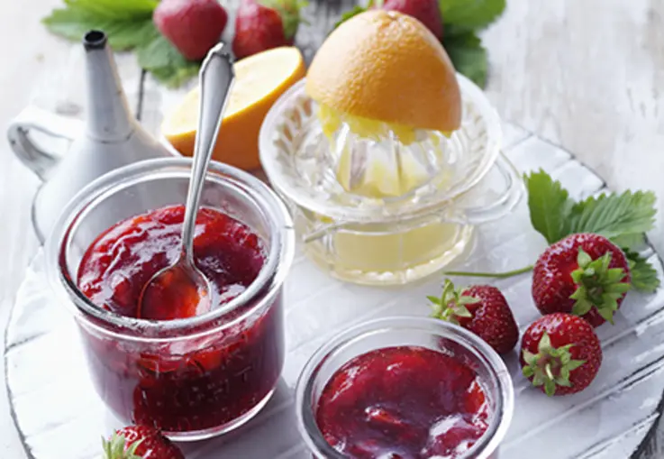 Glas mit dunkelroter Erdbeer‑Orangen‑Konfitüre, daneben frisch gepresste Orange und Erdbeeren