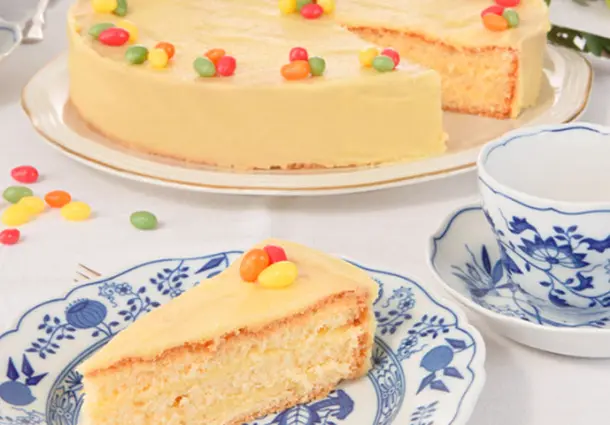 Eine gelb glasierte Ostertorte mit Zitronenbuttercreme, dekoriert mit bunten Zucker-Eiern; ein Stück liegt auf einem blau-weißen Teller