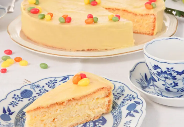Eine gelb glasierte Ostertorte mit Zitronenbuttercreme, dekoriert mit bunten Zucker-Eiern; ein Stück liegt auf einem blau-weißen Teller