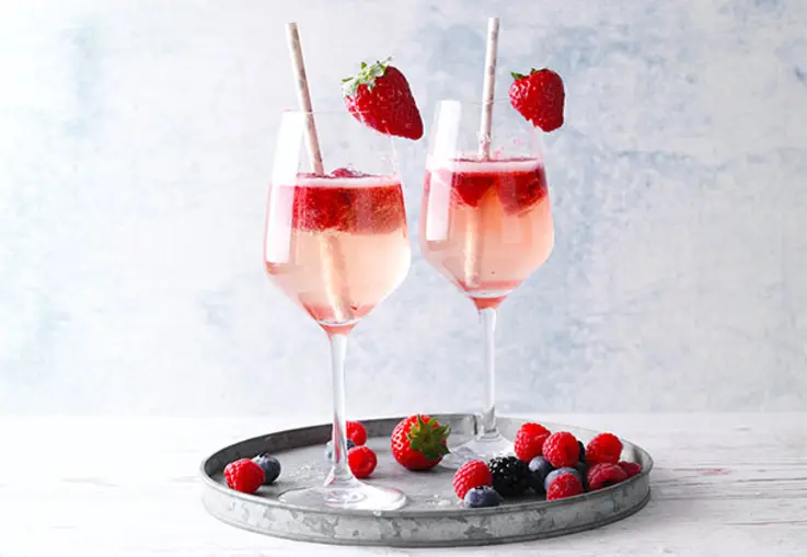 Ein Glas prickelnder Aperol-Spritz mit selbstgemachten Beeren-Eiswürfeln und frischen, roten Beeren schimmert auf einem Holztisch