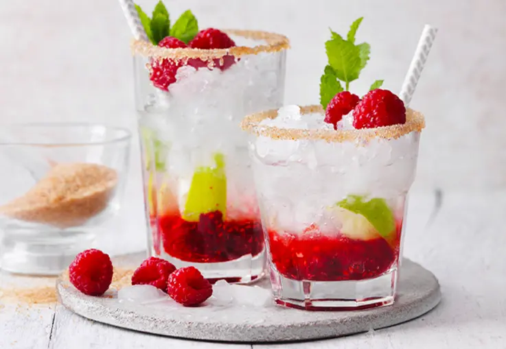 Ein Glas voller crushed Ice enthält zerdrückte Himbeeren und Limettenstücke; der Zuckerrand glitzert und frische Melissenblätter runden den Drink ab