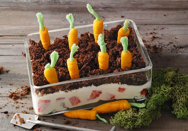 Ein Glasdessert mit Erdbeer‑Mascarpone‑Creme, darüber eine Schicht Schokobrösel und Marzipankarotten, die wie ein Hochbeet gepflanzt sind