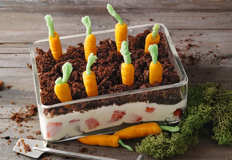 Ein Glasdessert mit Erdbeer‑Mascarpone‑Creme, darüber eine Schicht Schokobrösel und Marzipankarotten, die wie ein Hochbeet gepflanzt sind