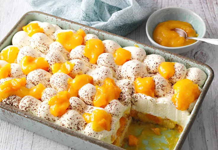 Ein rechteckiges Frucht‑Tiramisu in einer Glasform mit Mangopüree, Sahnecreme und Kakaopulver, daneben eine kleine Schüssel Mangopüree