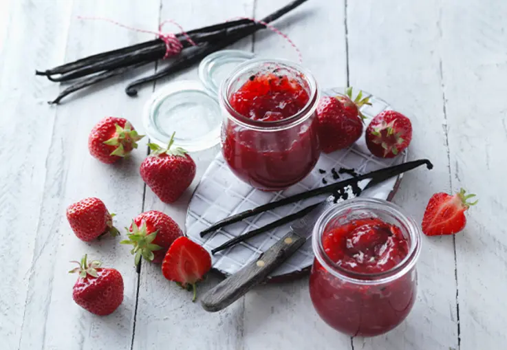 Kleine Gläser mit roter Konfitüre, umgeben von Vanilleschoten und frischen Erdbeeren