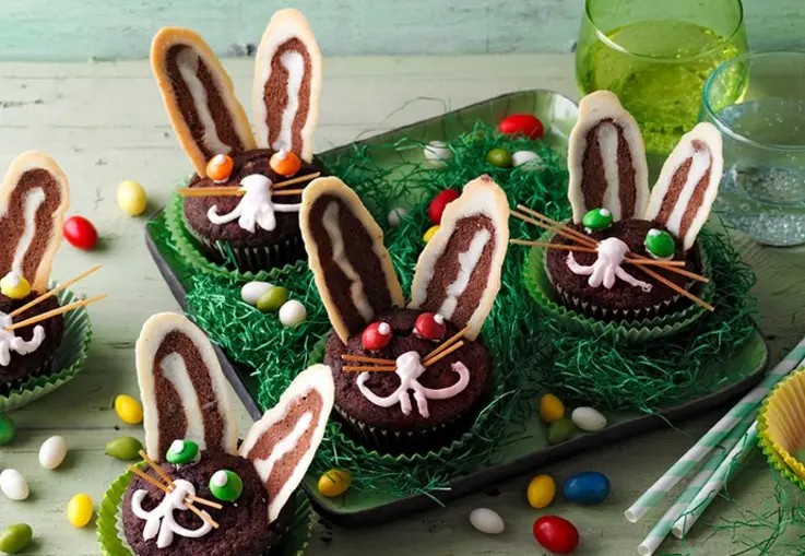 Schokoladige Muffins mit Möhren-Kakaoteig, verziert als Osterhasen mit langen zweifarbigen Ohren, rosa Zuckergussgesichtern und Schokolinsenaugen