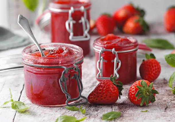 Ein Glas Erdbeer-Limetten-Konfitüre steht mit frischen Erdbeeren und Basilikumblättern auf einer Holzplatte