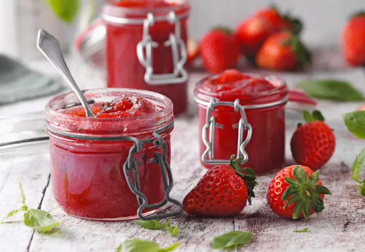 Ein Glas Erdbeer-Limetten-Konfitüre steht mit frischen Erdbeeren und Basilikumblättern auf einer Holzplatte