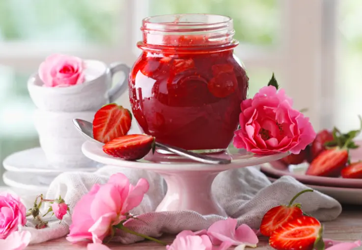 Ein großes Glas mit samtig roter Erdbeerkonfitüre, durchzogen von feinen Rosenblättern und glänzender Oberfläche.